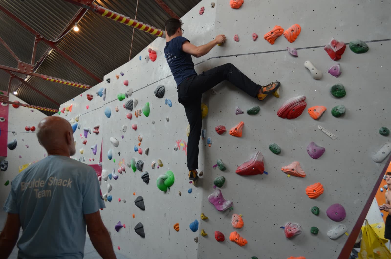 A climber on the wall with a coach watching from the mats